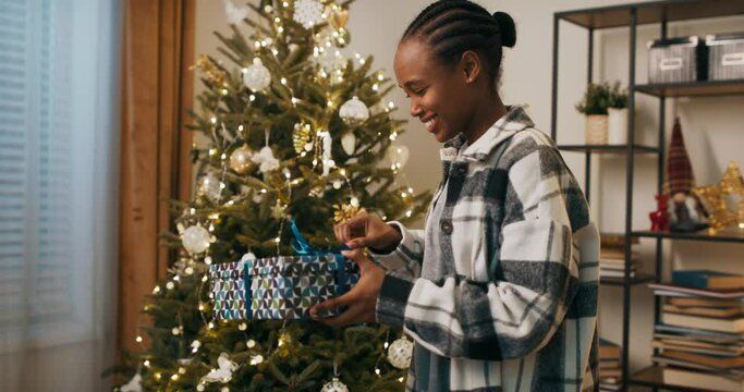 An African-looking student stands by Christmas tree and unpacks gift. She unties the blue ribbon and opens the box. The girl received warm black hat and hugged it. She is smiling and happy.