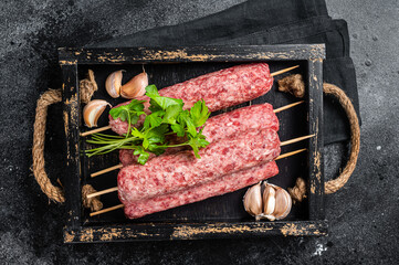 Fresh raw beef meat kebabs sausages on skewers in wooden tray. Black background. Top view