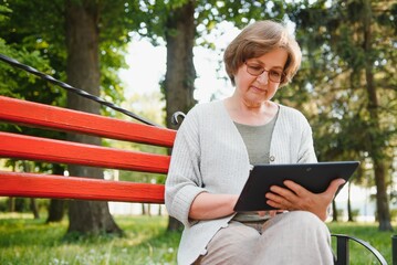 Fototapeta premium Elegant elderly woman in the shirt is sitting on the bench in a park on a warm day