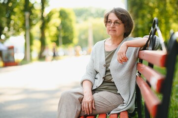 Fototapeta na wymiar Elegant elderly woman in the shirt is sitting on the bench in a park on a warm day