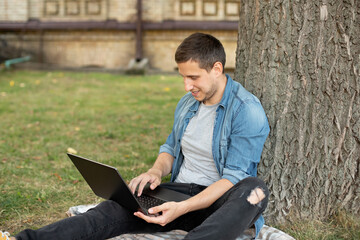 Smilling Young man with laptop rest in grass at university campus. Business man sit on lawn work outdoors on computer in a park. Student man on lesson with laptop. education and remote working concept