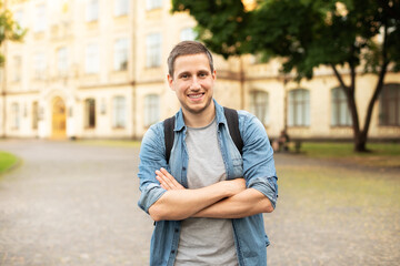 Fototapeta premium Successful student is standing with backpack near campus on the university background. Smiling young man with bag standing in park. Education, university, college, studying, course concept 