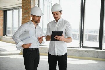 A front view of two smart architects with white helmets reviewing blueprints at a construction site on a bright sunny day