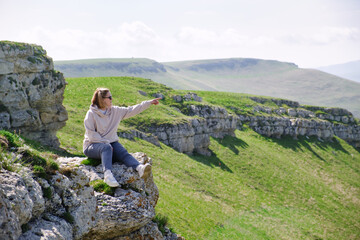 A woman tourist sits on the rock and pointing into the distance by her hand. Female tourist in mountains.