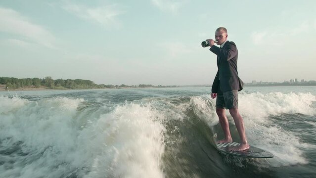 A happy surfer jumping on a wakeboard in a suit with a bottle of champagne. An experienced wakeboarder sprays water drops into the camera.