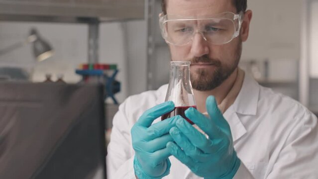 Chest up slowmo of Caucasian male chemist in lab coat and protective goggles taking smell of red liquid substance inside glass flask, conducting experiment in laboratory