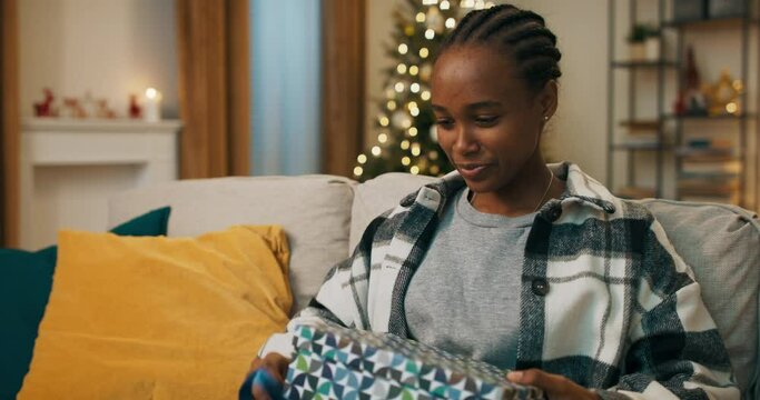 Winter evening. An African-looking girl sits on sofa in the living room and unpacks gift. She looks forward to opening the box and sees horrible black hat she doesn't like. The girl was upset.