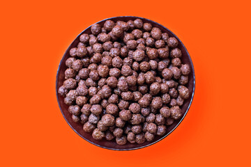 Chocolate cornflakes in a bowl on an orange isolated background in the center of the image. Flakes with chocolate flavor, close-up top view. American-style energy carbohydrate breakfast