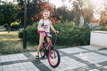 A child girl with pigtails rides in the park on a red bicycle and is dressed in summer clothes. A...