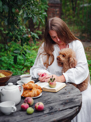 middle aged caucasian woman in white dress enjoying morning coffee in apple orchard sitting at wooden table with her pet poodle