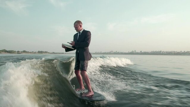A surfer jumping on a wakeboard in a suit with a book in his hands takes photos of the pages of the book with his phone. An experienced wakeboarder sprays water drops into the camera.