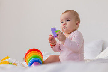 baby in a pink bodysuit plays with an educational multicolored pyramid while sitting in bed