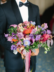 The groom in a black suit and bow tie holds the bride's wedding bouquet of exotic flowers.