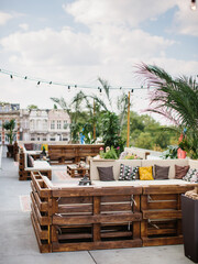 Furniture from wooden pallets with colorful cushions on the roof terrace. Palm trees in wicker flowerpots, garlands with light bulbs.