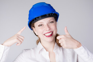 Young female engineer is pointing to her hart hat and shows thumbs-up