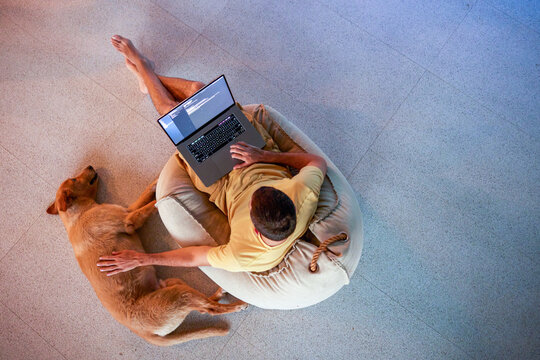 Overhead view of man using laptop computer, sitting at home on bean bag chair next to his big golden retriever dog