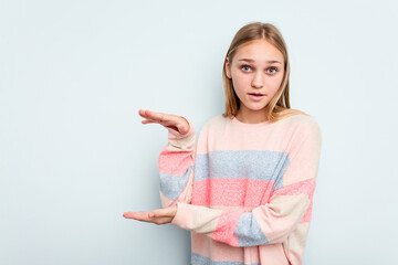 Young caucasian girl isolated on blue background shocked and amazed holding a copy space between hands.