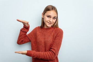 Young caucasian girl isolated on blue background holding something little with forefingers, smiling and confident.