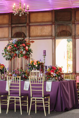 Tropical style purple banquet table wedding decor. Purple tablecloth, exotic flowers, white napkins, dishes, cutlery, and purple glasses. In the foreground are beige chairs. At the top is a classic 