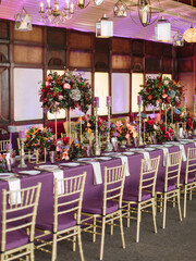 Tropical style purple banquet table wedding decor. Purple tablecloth, exotic flowers, white napkins, dishes, cutlery, and purple glasses. In the foreground are beige chairs.