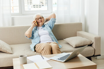 a happy, relaxed elderly lady works from home sitting on a cozy sofa in a bright interior and talking on the phone with her hand behind her head