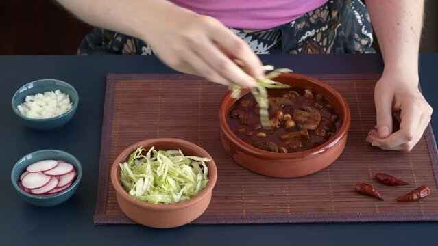 Decorating vegan Pozole Rojo with shredded ice lettuce, radish and white onion. Mexican dish with hominy, shredded jackfruit, button mushrooms and dried chilies: guaillo, ancho, pasilla.