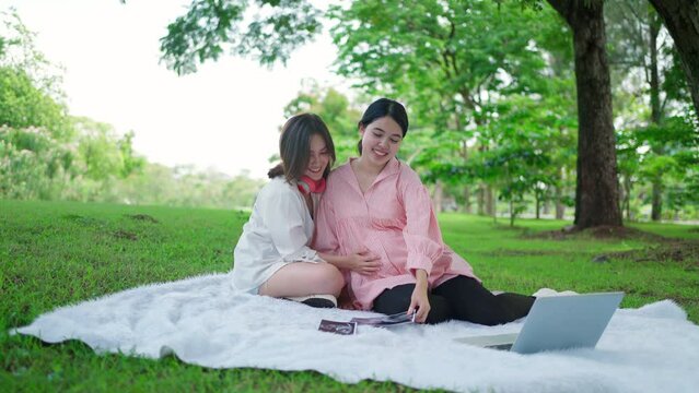 Asia beautiful pregnant woman sitting in the public garden with her buddy and stroking her belly in the sunny day. Looking at photo of ultrasound scan of her baby. Communicate with an embryo.