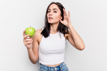 Young caucasian woman holding an apple isolated on white background trying to listening a gossip.