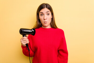 Young caucasian woman holding hairdryer isolated on yellow background shrugs shoulders and open eyes confused.