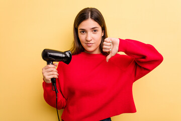 Young caucasian woman holding hairdryer isolated on yellow background showing a dislike gesture, thumbs down. Disagreement concept.