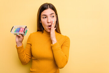Young caucasian woman holding a batteries box isolated on yellow background is saying a secret hot braking news and looking aside