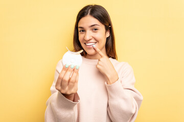 Young caucasian woman holding teeth whitener isolated on yellow background