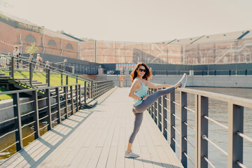 Glad brunette woman leans at fence stretches legs has perfect figure dressed in activewear poses outdoor demonstrates her flexibility leads sporty lifestyle warms up before fitness training.