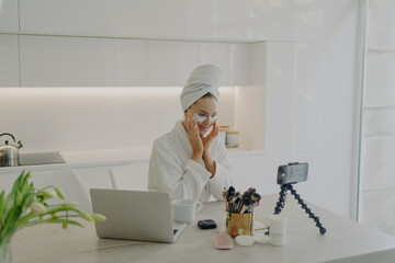 Female beauty blogger in bathrobe and towel on head recording video on smartphone for her vlog