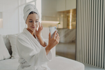 Young beautiful woman applying cosmetic cream while relaxing on sofa after taking shower at home