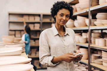 Confident young ceramist using a smartphoe in her shop