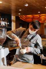 A masked barista prepares an exquisite delicious coffee at the bar in a coffee shop. The work of restaurants and cafes during the pandemic.