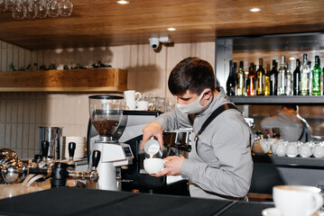 A masked barista prepares an exquisite delicious coffee at the bar in a coffee shop. The work of restaurants and cafes during the pandemic.