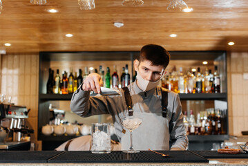 A stylish bartender in a mask and uniform during a pandemic is preparing cocktails at a party. The work of restaurants and cafes during the pandemic.
