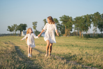 Naklejka na ściany i meble A mother and daughter run into the field by the hand. Happy childhood with my mother. Daughter and mother in a white dress in the field. Psychology of parents' relations with children