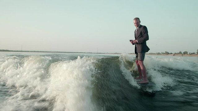 A surfer jumping on a wakeboard in a suit with a phone in his hands. An experienced wakeboarder sprays water drops into the camera.