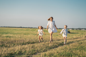 Naklejka na ściany i meble World Women's Day. A happy mother with her daughters runs into the field. Psychology of the relationship between mother and daughter. Life in a modern village