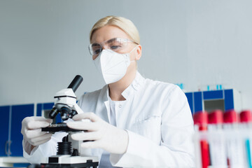 Scientist in goggles and latex gloves using microscope near blurred test tubes in lab.