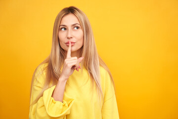 Shh blonde woman asks to keep quiet or secret keeps forefinger on lips isolated on yellow background