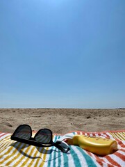 Vertical image of a beach towel, sunglasses and a spray of sunscreen with the sand of the beach in a lonely background. Concept of summer vacation at the sea.