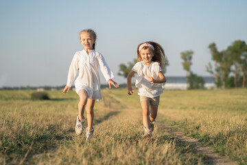Two beautiful girls happily run into the field among the grass. Childhood in the village with children. Happy kids running into the field