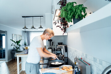 A young man is cooking cheese pancakes in a modern light kitchen at home. Man preparing pancakes for frying on the pan. Housework and home duties. Culinary hobby. Selective focus