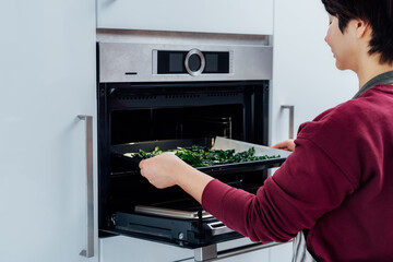 Woman putting baking sheet with teared curly green kale leaves into oven the modern kitchen....