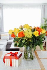 Yellow and orange roses in a vase and gift box with red ribbon on kitchen counter table with open space living room background. Gift for Thanksgiving day, birthday, Mother's or Valentine's day.