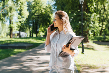 Cheerful female student in eyeglasses with cute smile calling on mobile phone and holding laptop while standing in park, looking away. Portrait of young woman freelancer using smartphone outdoors